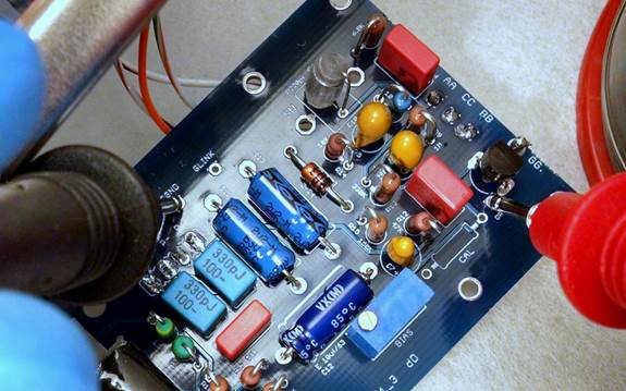 Two multimeter probes on the D-87 PCB at the FET drain pin and chassis ground. Blue trim pot visible center-right.